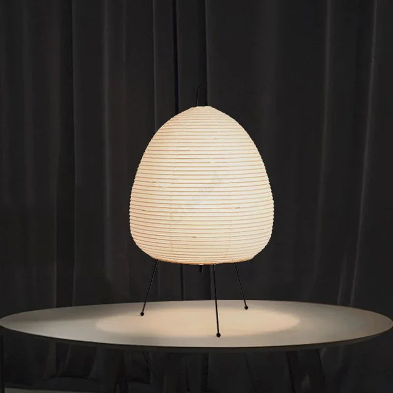 Modern paper lantern table lamp with tripod legs on white round table, dark curtain backdrop